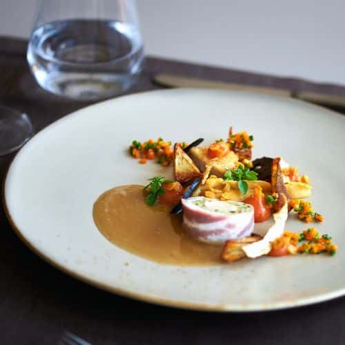 Ballotin de volaille farci entouré de lard, servi avec légumes racines, champignons et jus corsé dans une assiette gastronomique